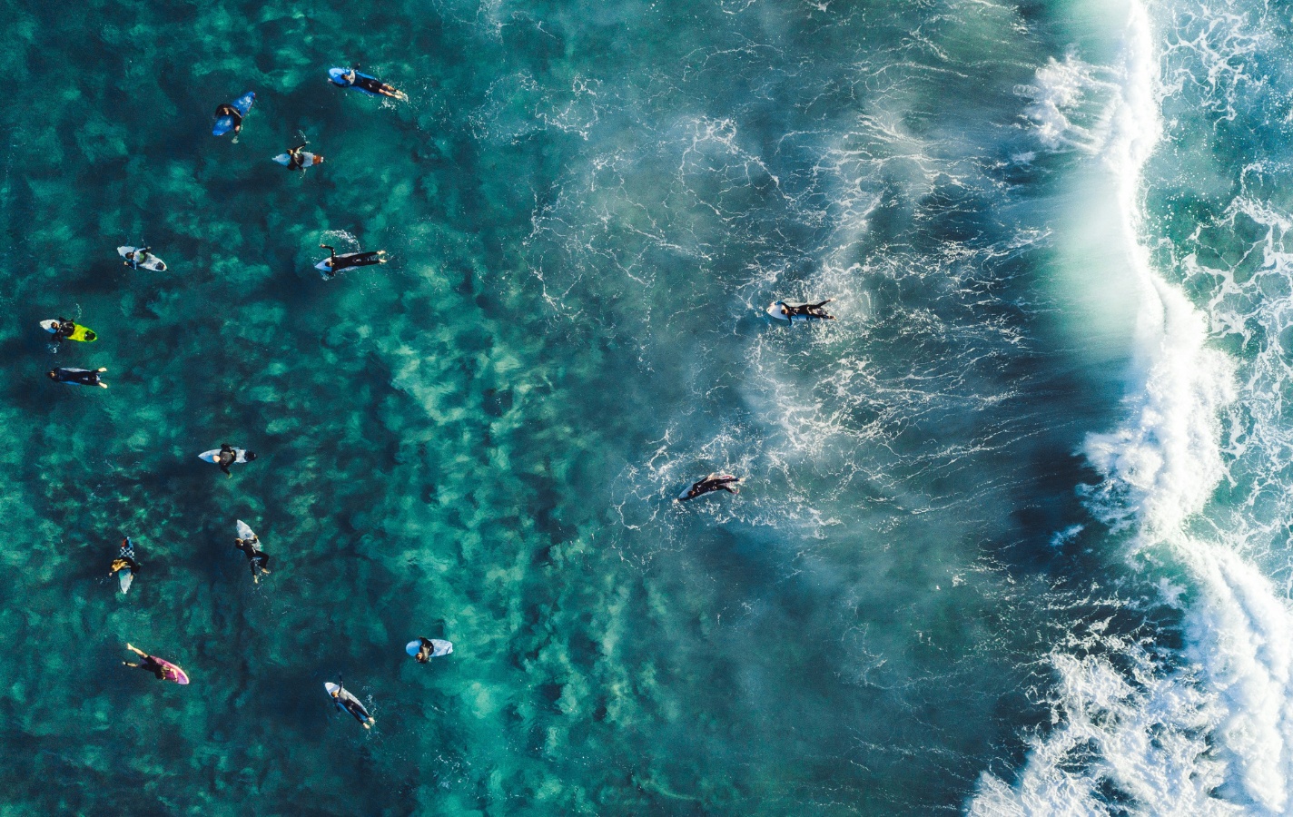 An aerial view of surfers paddling in crystal-clear reef waters, just beyond the break — a dynamic scene of community, energy, and the call of the wave.