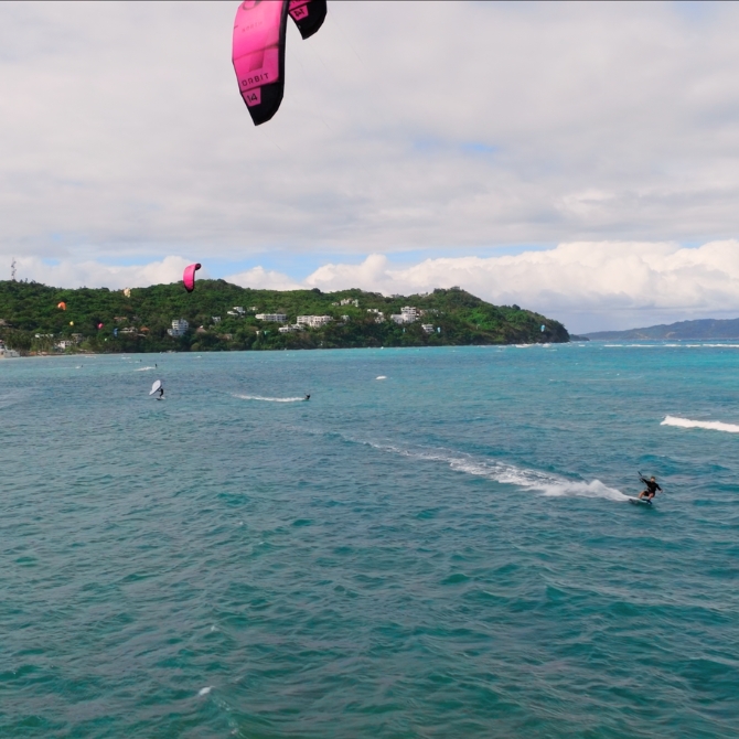 Two kite surfers ride side by side on turquoise waters off a lush island coastline, capturing the wild freedom and camaraderie that ignited the outriderx story.